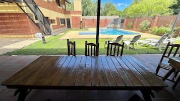 A wooden patio table with four chairs, a pool, and a brick building in the background.