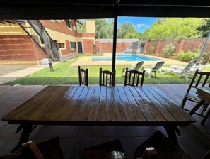 Een houten tuintafel met vier stoelen, een zwembad en een bakstenen gebouw op de achtergrond.