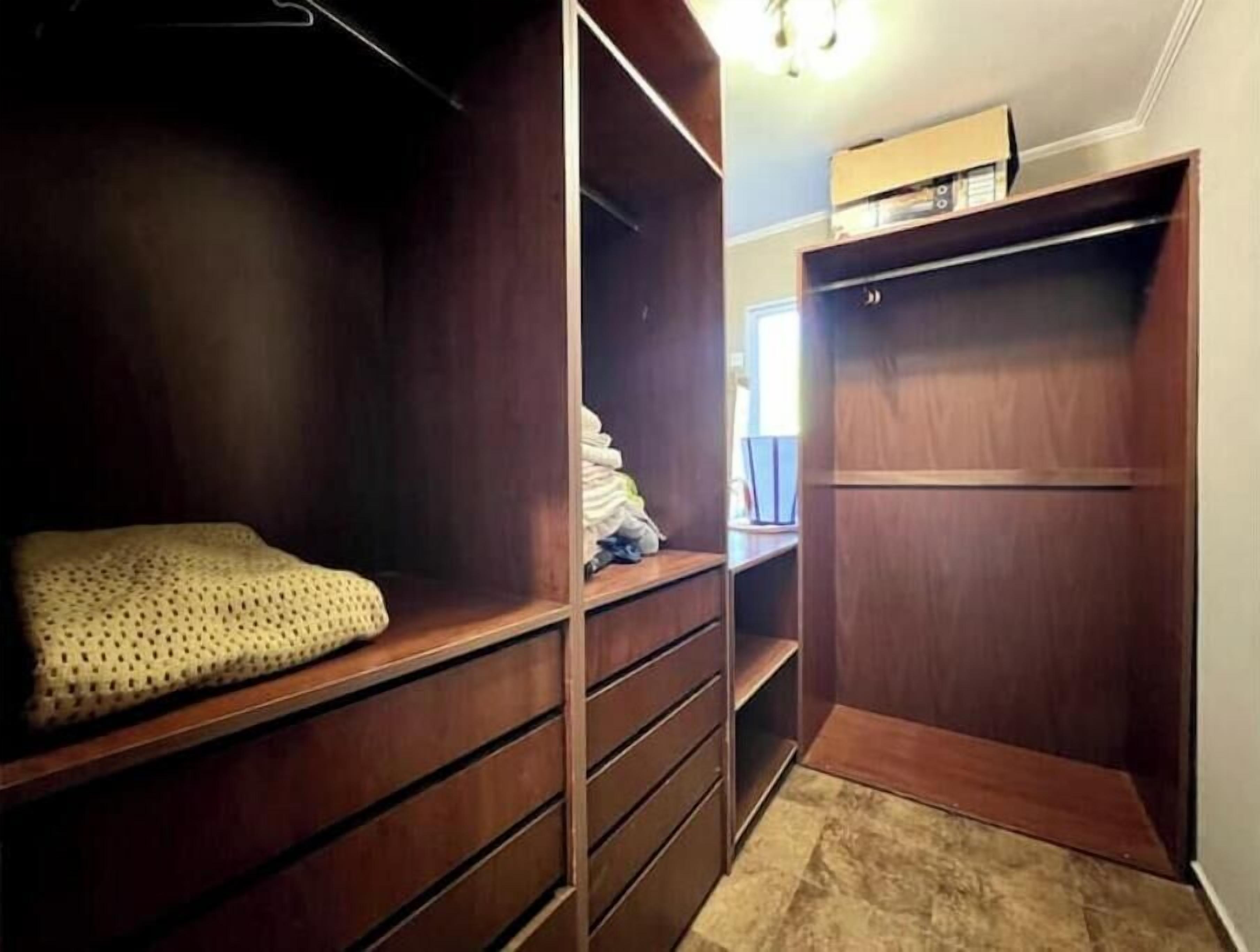 Un dressing accessible sans porte, doté d’étagères et de tiroirs en bois, un petit coin salon avec un coussin à motifs et une boîte posée sur le dessus du dressing.