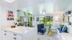 A modern kitchen with white cabinetry and a central island, a cozy seating area with a blue sofa and a glass coffee table, and a large window offering a view of the outdoors.