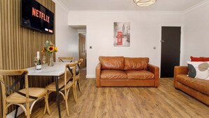 A modern living room with a leather sofa, wooden dining table, and chairs. A flat-screen TV mounted on the wall displays the Netflix logo. - Walpole Retreat - Hot Tub, Games Room, Sleeps 4 (York)