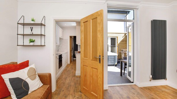 A modern living room with a wooden door, a sofa with cushions, a metal shelf with plants, and a view into the kitchen and dining area. - Walpole Retreat - Hot Tub, Games Room, Sleeps 4 (York)
