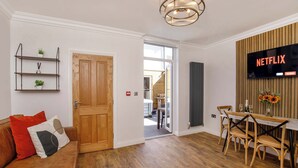 A modern living room with a wooden door, a dining area with a table and chairs, a TV mounted on a wooden panel, and a bookshelf with decorative items. - Walpole Retreat - Hot Tub, Games Room, Sleeps 4 (York)
