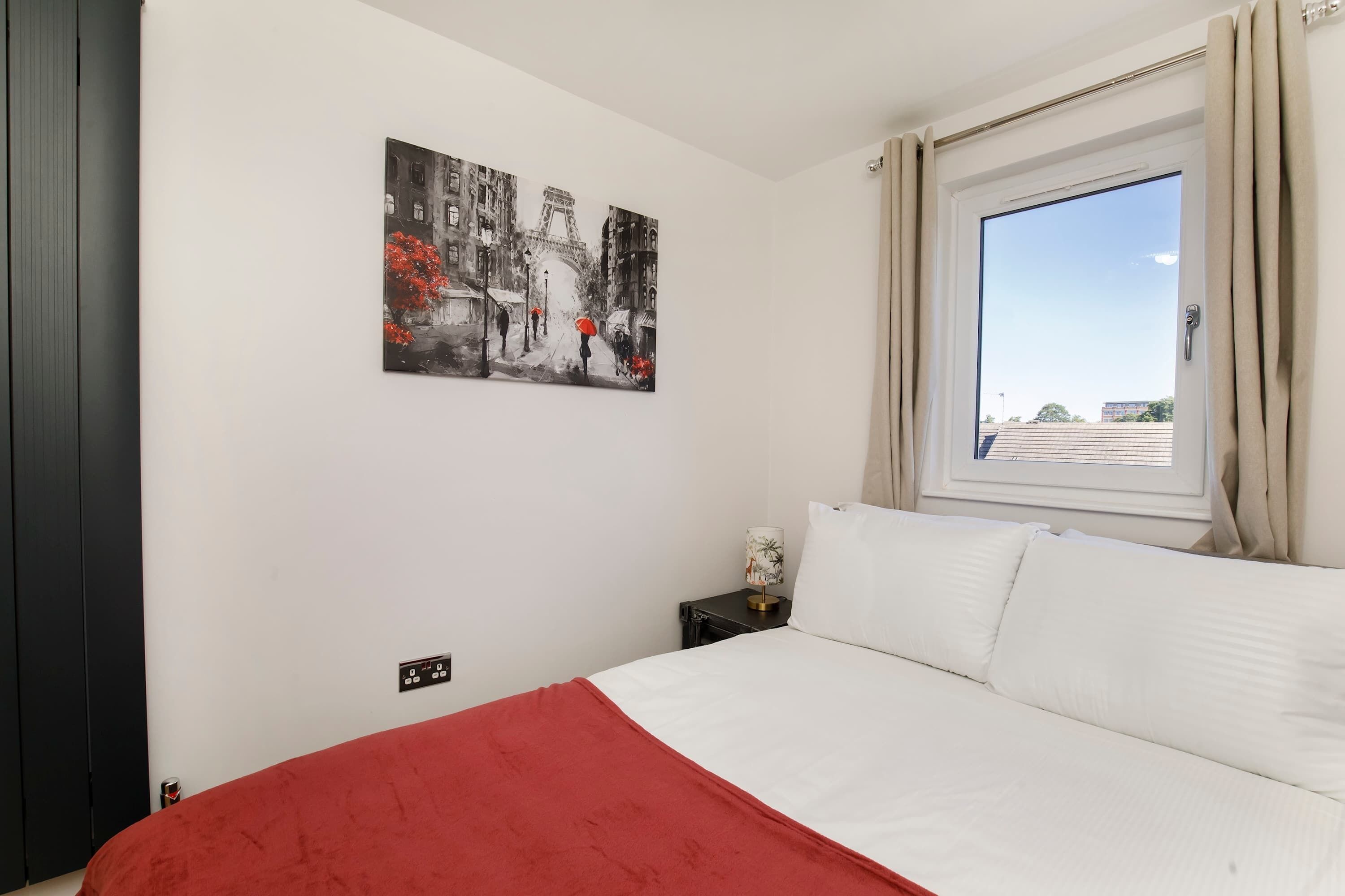 A bedroom with a bed, a nightstand, a lamp, a window with curtains, and a painting on the wall.