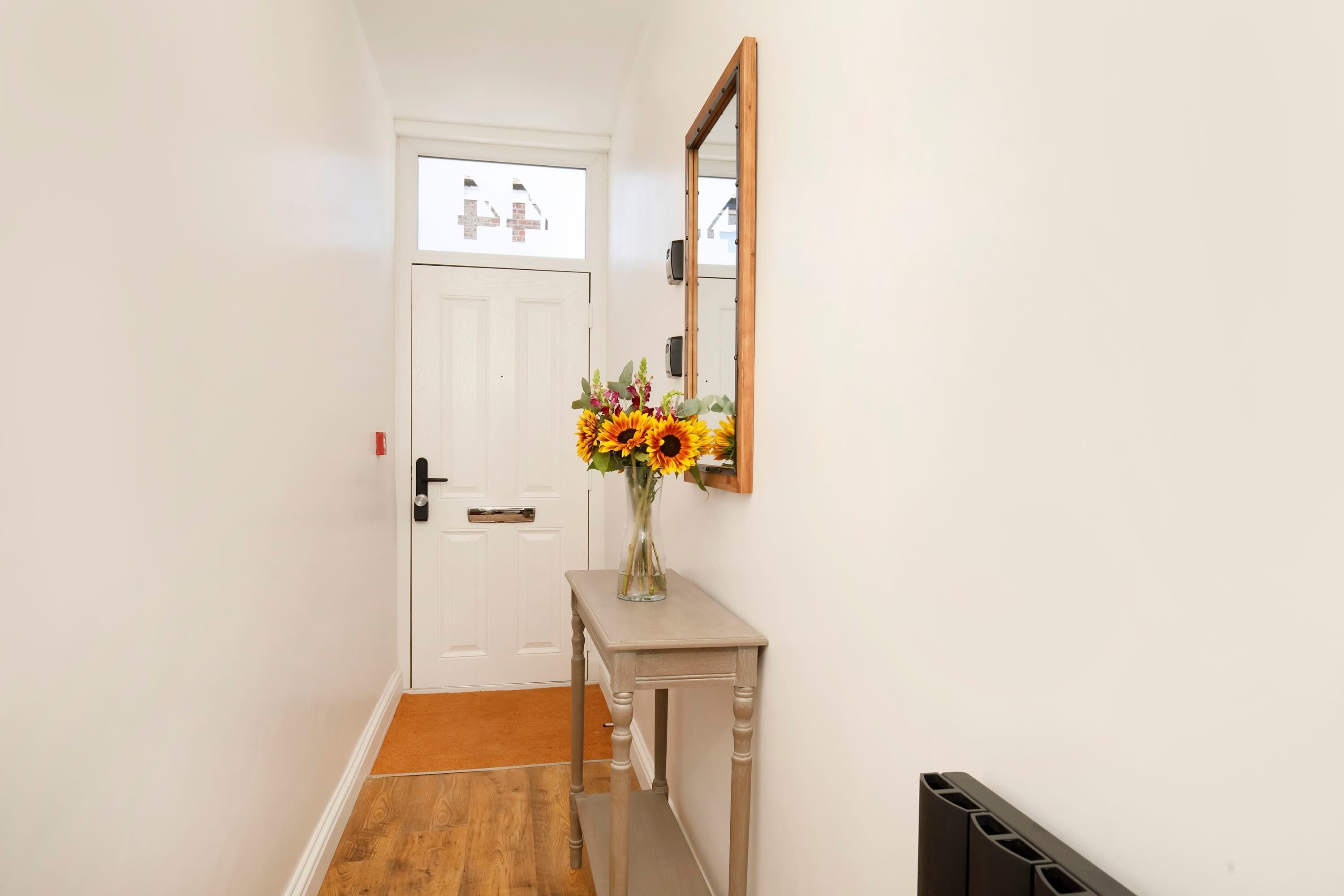 Un pasillo con una puerta blanca, piso de madera, una pequeña mesa con un florero y un espejo en la pared.