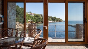 A glass-enclosed patio with wicker furniture, a stone wall, and a view of a coastal area with a body of water and greenery. - Porto Castello Villas (Skiathos)