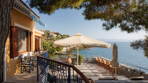 A terrace with outdoor seating, a view of the sea, and a building with a balcony. - Porto Castello Villas (Skiathos)
