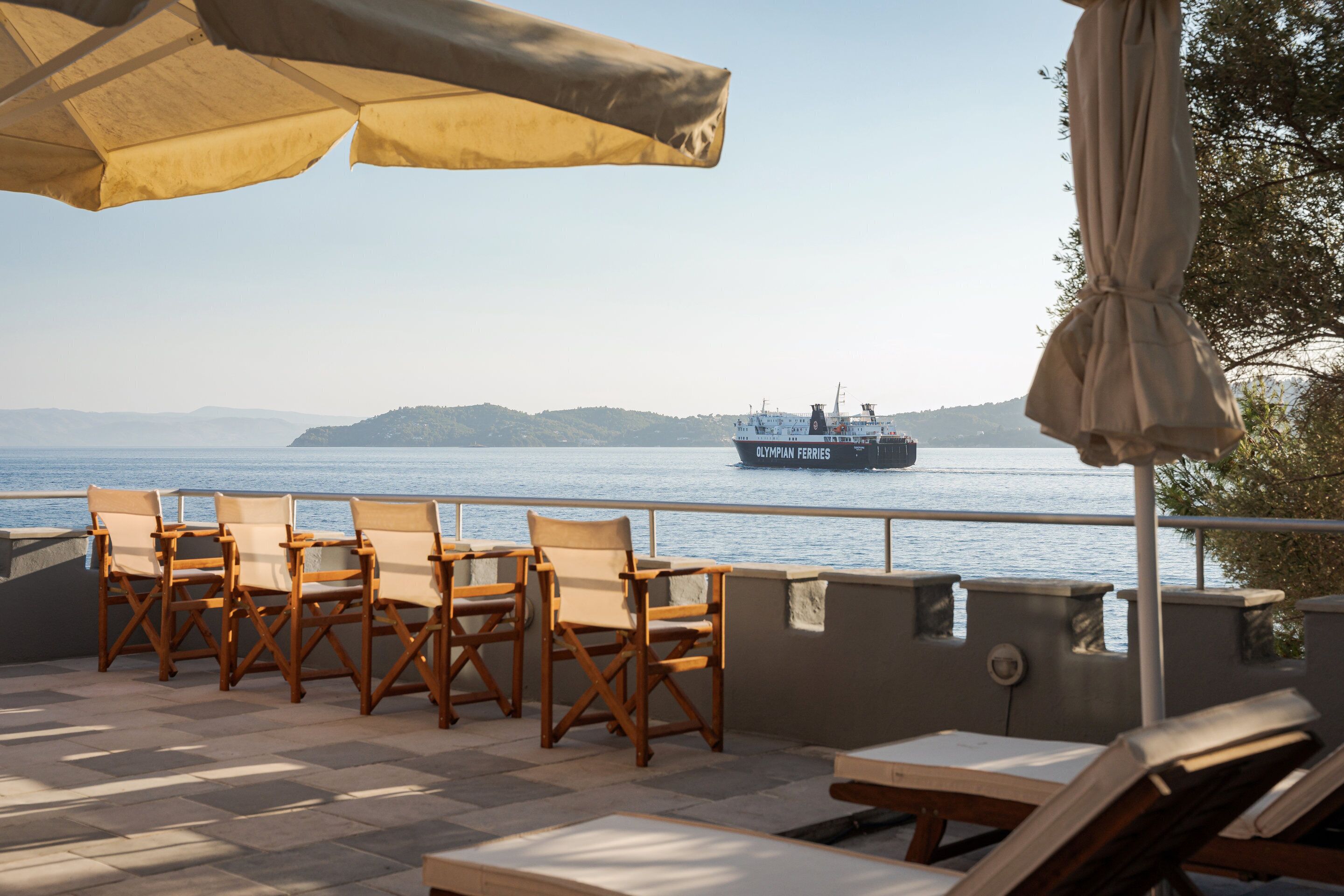 A terrace with wooden chairs and tables, overlooking a body of water with a ship in the distance.