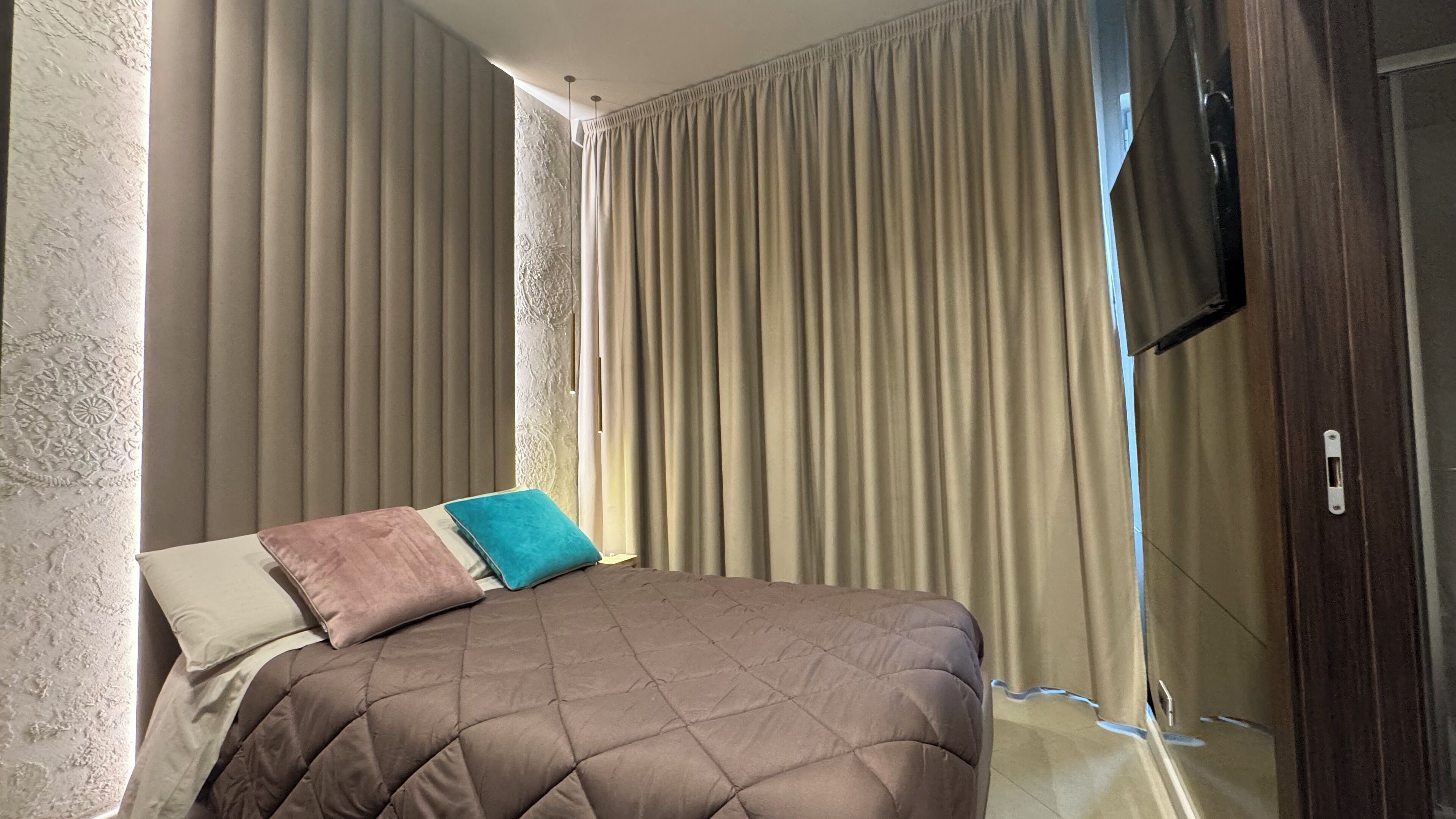 A bedroom with a bed, beige curtains, and a TV mounted on the wall.