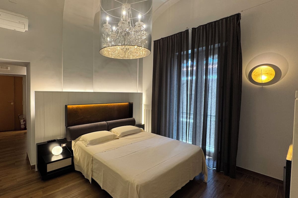 Une chambre moderne avec un grand lit, une table de chevet avec une lampe, un lustre et une vue sur l’extérieur à travers des rideaux légers.
