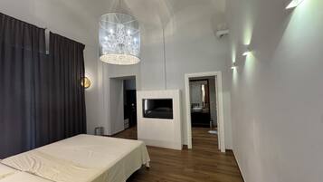 Ein modernes Schlafzimmer mit einem Bett, einem an der Wand montierten Fernseher, einer Kristallleuchtre und einem durch eine offene Tür einsehbaren Badezimmer.