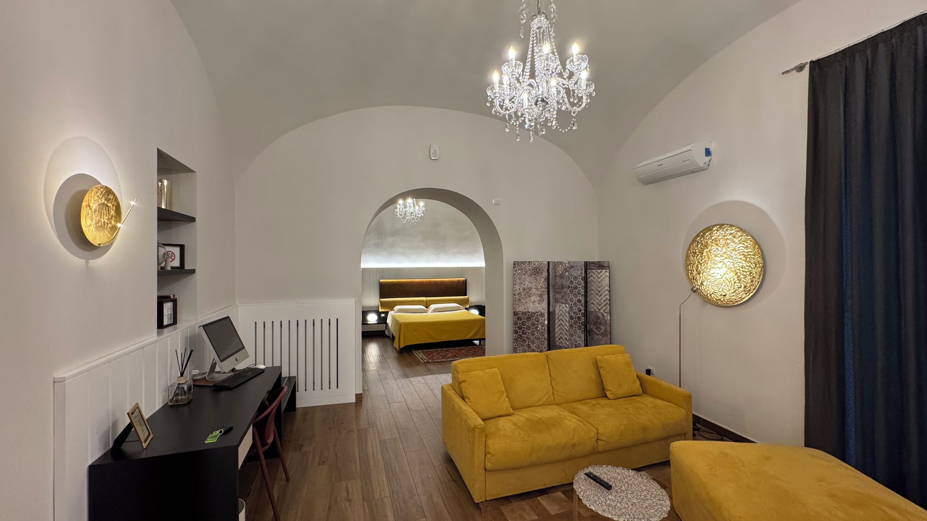 A modern living room with a yellow sofa, a black desk with a computer, a chandelier, and a bed in the background.