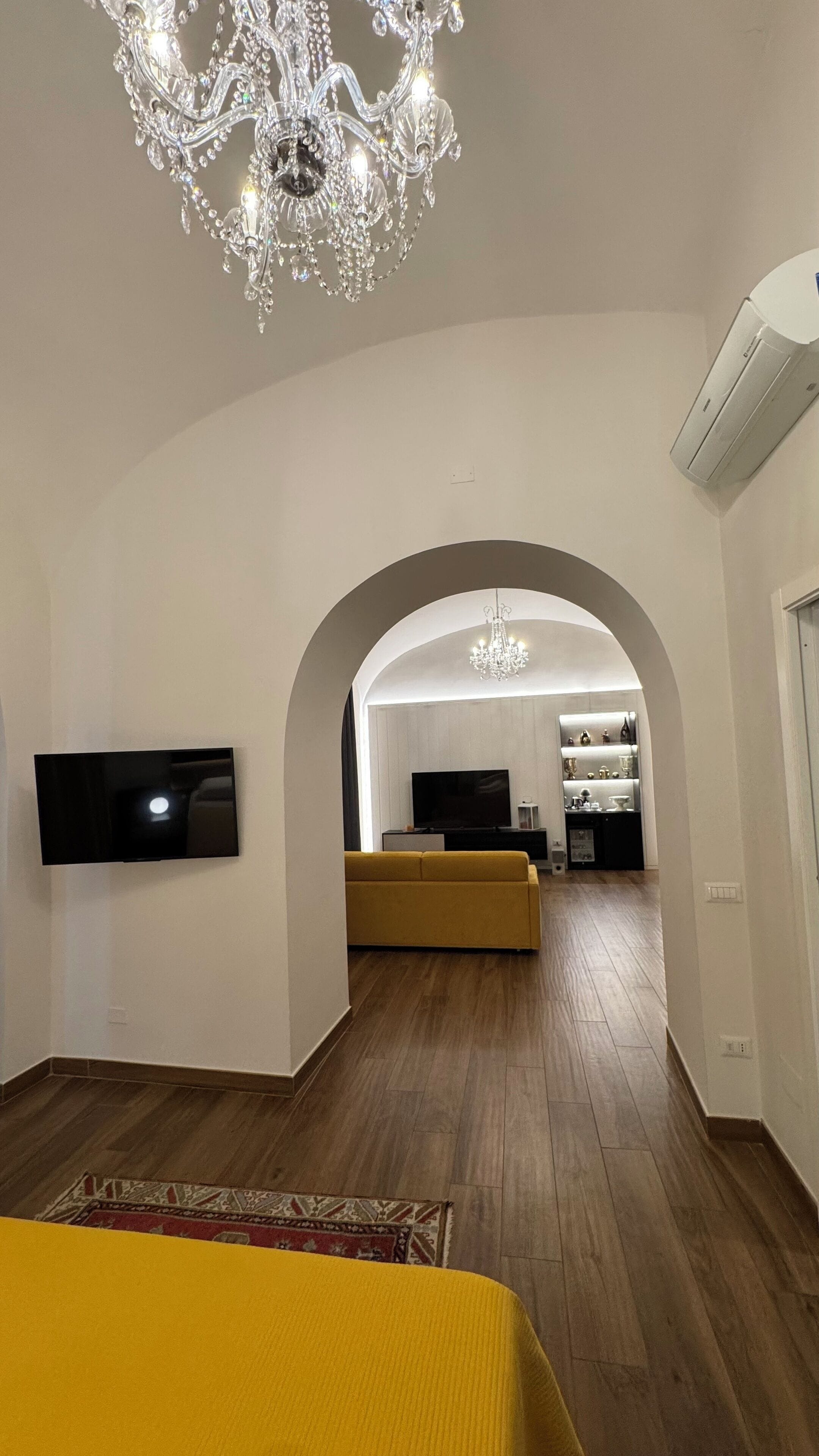 Ein modernes Wohnzimmer mit einem großen Kronleuchter, einem Flachbildfernseher, einem gelben Sofa und Holzboden.
