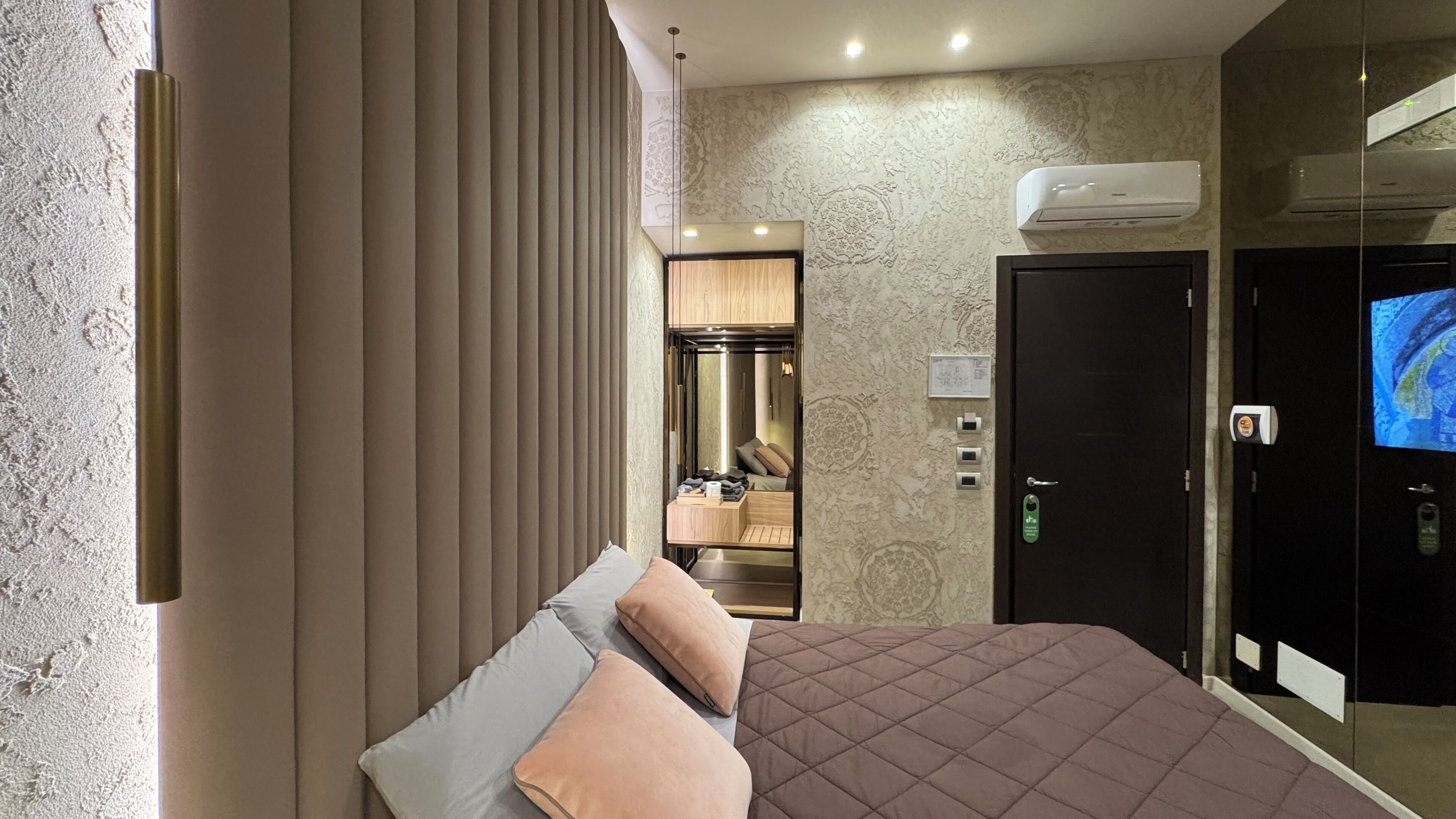 A hotel room with a bed, a TV, an air conditioning unit, and a door with a lock and keycard reader.