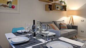 Un comedor con una mesa preparada para cuatro personas, con copas de vino, platos y cubiertos. Se observa un sofá y una lámpara al fondo.