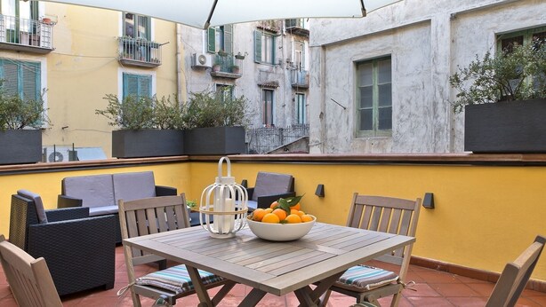 Un patio con mesa, sillas, un cuenco de fruta y plantas en macetas.