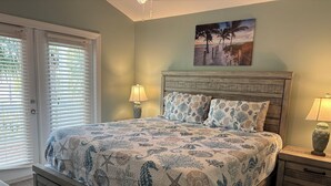 Une chambre à coucher comprenant un lit, deux tables de chevet, une tête de lit en bois, un tableau sur le thème de la plage et une porte avec des stores.