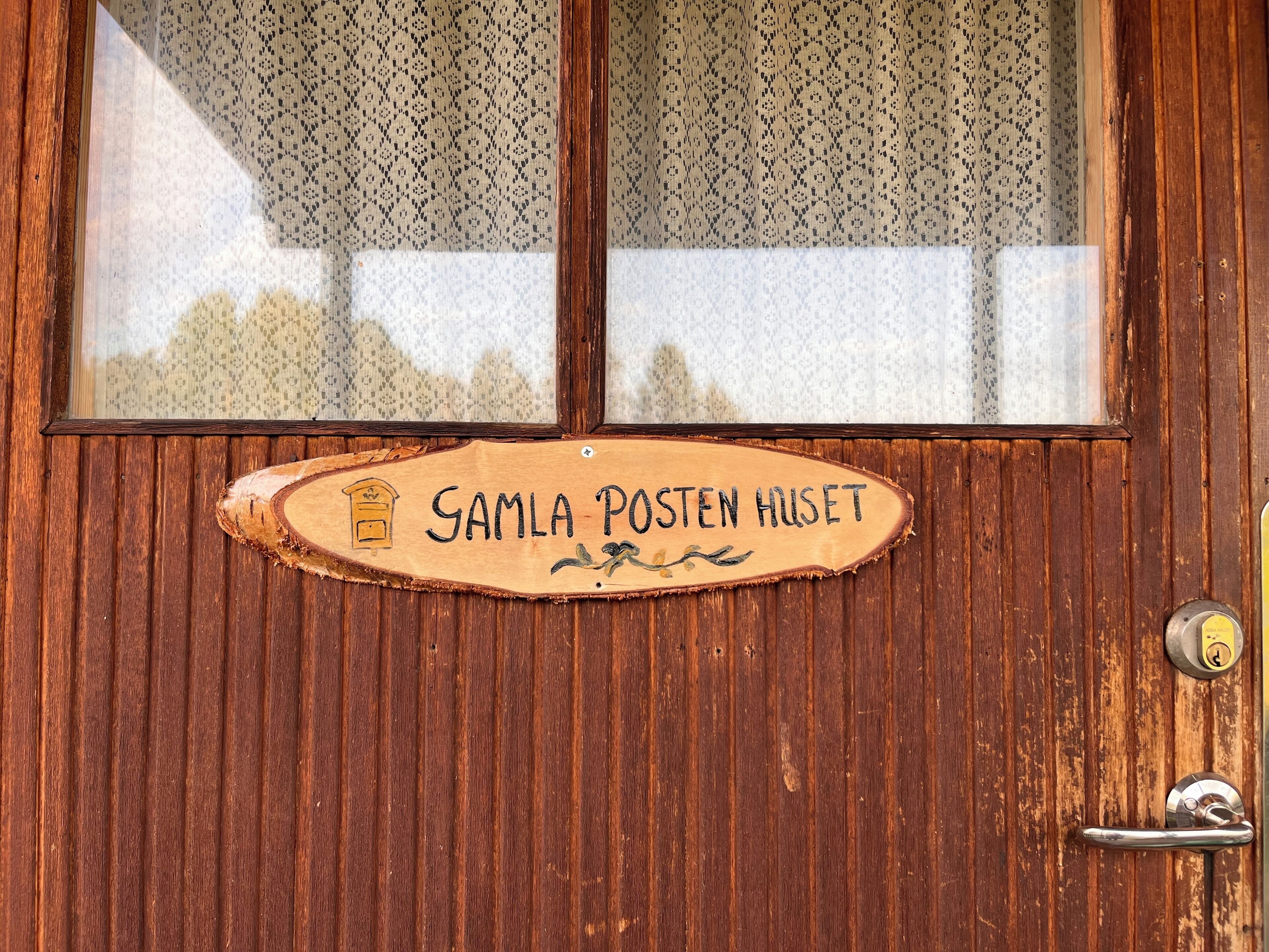 Une porte en bois avec une plaque indiquant « Samla Posten Huset » et une fenêtre ornée de rideaux à motifs.