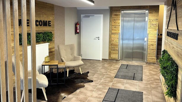 Eine moderne Lobby mit einer Rezeption, einem Feuerlöscher, einer weißen Tür, einem Aufzug und einer Empfangswand mit dem Schriftzug 'WELCOME'.