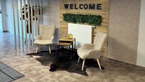 Ein moderner Empfangsbereich mit einem 'WELCOME'-Schild, zwei Stühlen, einem kleinen Tisch, einer Zimmerpflanze und einem gemusterten Teppich.