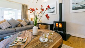 A cozy living room with a sofa, a wooden coffee table, a vase with flowers, a magazine, and a wood-burning stove.