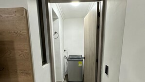 A narrow hallway with a wooden cabinet on the left, a laundry machine in the center, and a white door on the right. - Loft studio apartment (Envigado)