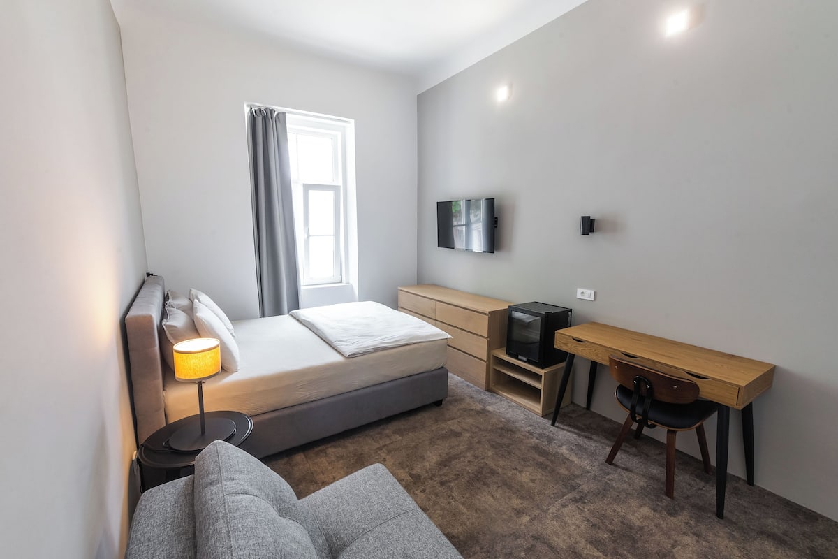 A modern bedroom with a bed, bedside table, lamp, TV, desk, chair, and a window with curtains.