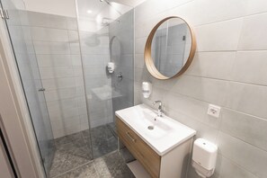 Un bany modern amb un box de dutxa de vidre, un lavabo blanc amb un moble de tocador de fusta i un mirall rodó a la paret.