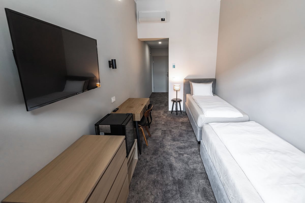 A compact hotel room with a flat-screen TV, a wooden desk, two single beds, and a small table with chairs.