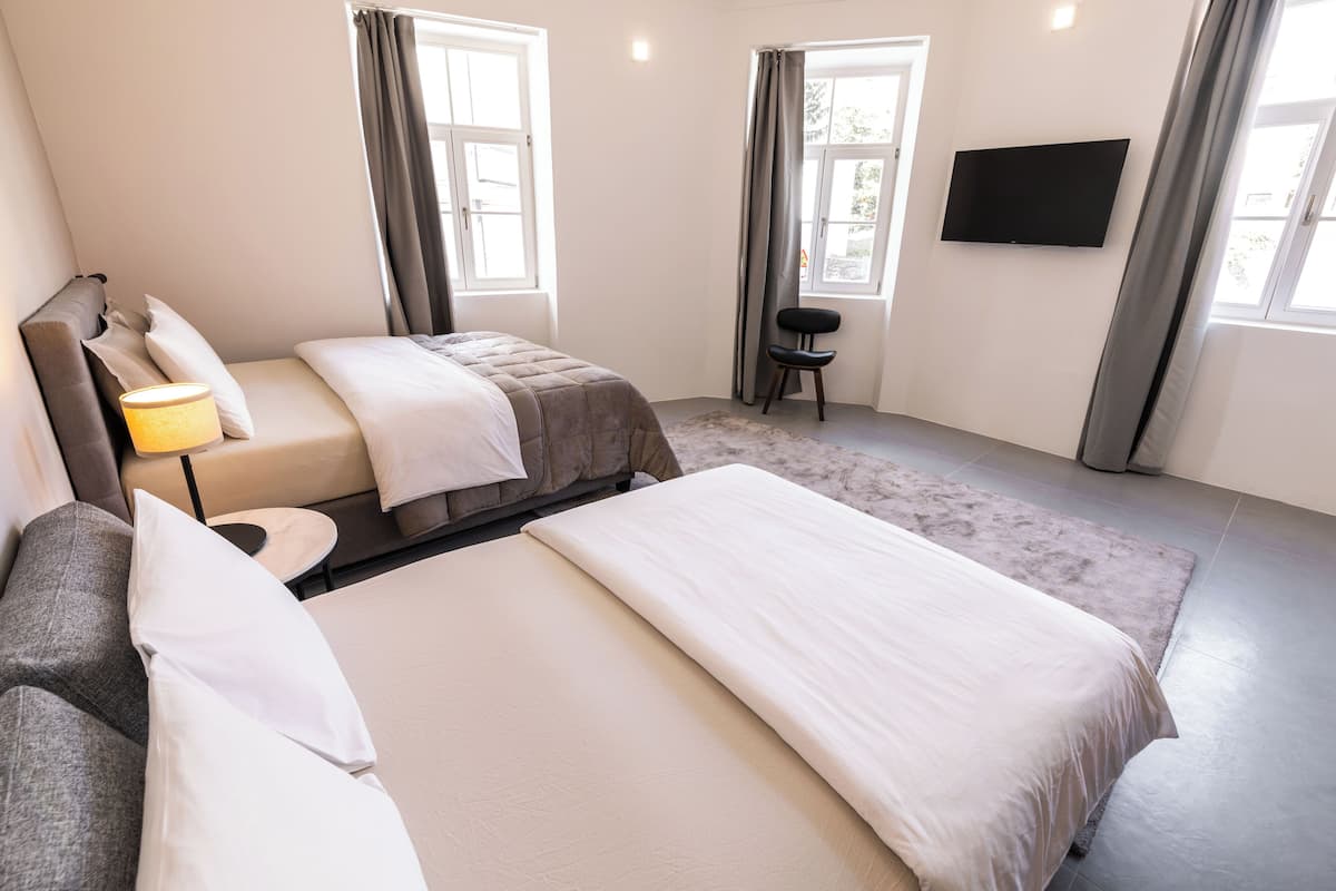 A hotel room with a bed, a sofa, a chair, a TV, and two windows with curtains.