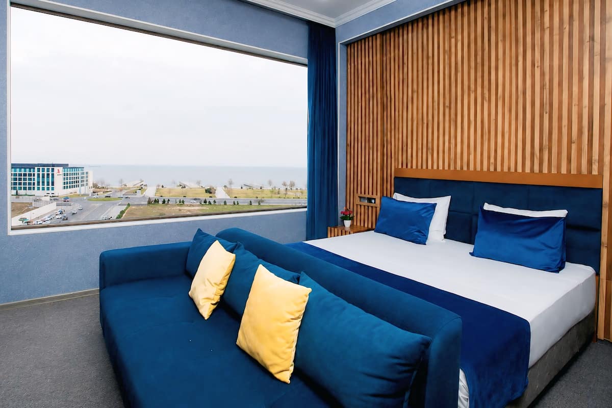 Una habitación de hotel moderna con un ventanal grande, una cama con ropa de cama azul y blanca, un sofá azul con almohadas amarillas y un panel de pared de madera.