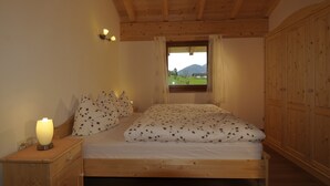 A wooden bed with a floral bedspread, a bedside lamp, a window with a view of a green landscape, and a wooden wardrobe. - Apartment 'Sunny' with Mountain View, Shared Pool and Wi-Fi (Ratschings - Mareit)
