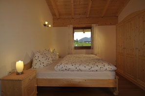 Un llit de fusta amb una colcha floral, una làmpada de nit, una finestra amb vista a un paisatge verd i un armari de fusta.