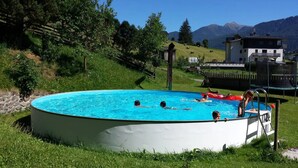 A round swimming pool surrounded by grass, with a few people swimming and a trampoline in the background. - Apartment 'Sunny' with Mountain View, Shared Pool and Wi-Fi (Ratschings - Mareit)