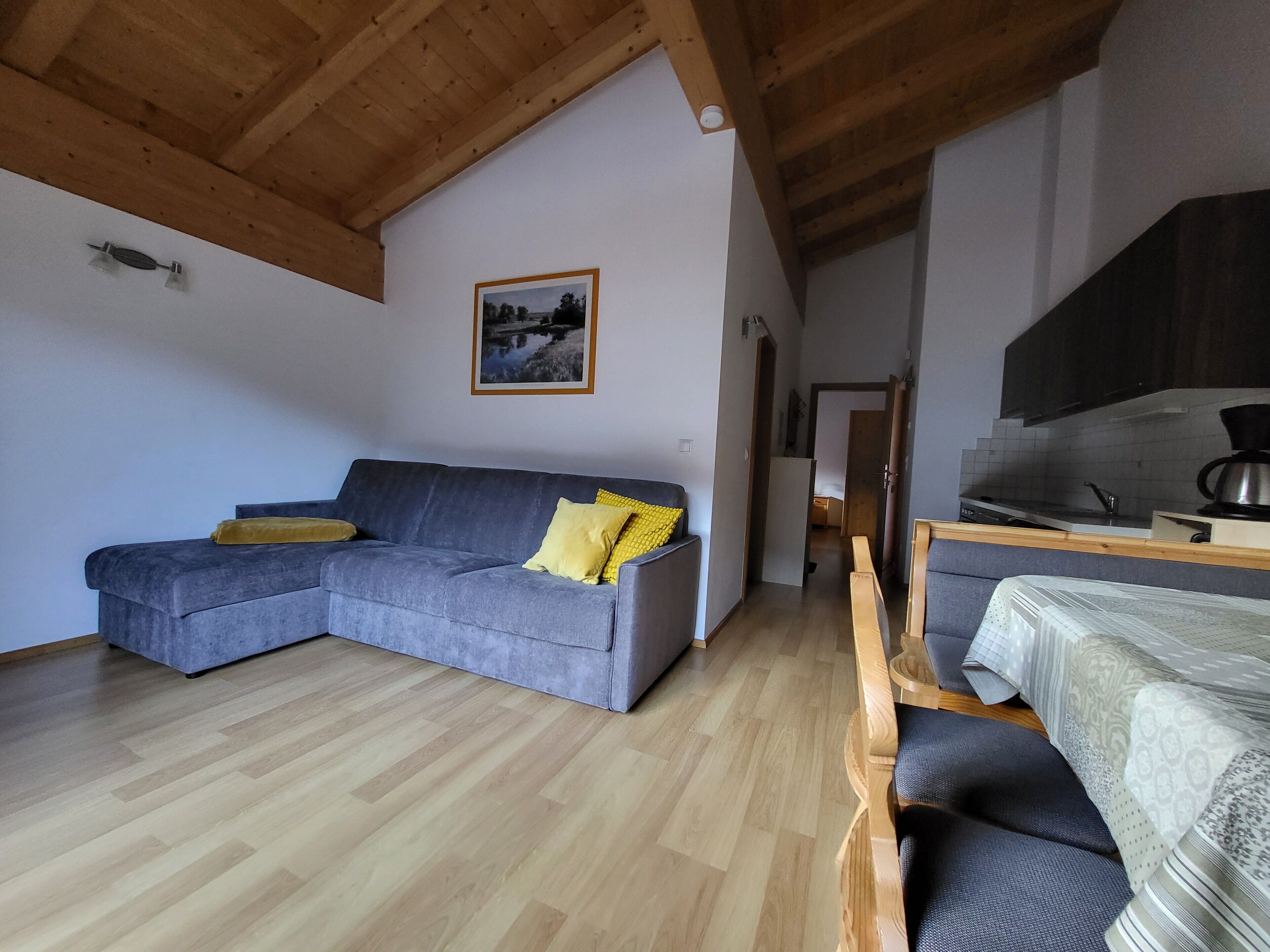A living area with a grey sectional sofa, wooden flooring, a kitchen with a stove, and a framed picture on the wall.