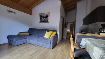 A living area with a grey sectional sofa, wooden flooring, a kitchen with a stove, and a framed picture on the wall.