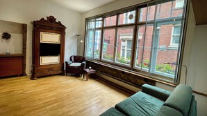 A room with a wooden floor, a green sofa, a wooden cabinet with a television, and large windows with a view of a brick building.