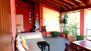 Une terrasse couverte avec un canapé, une table et des chaises, entourée de plantes en pot et d’un barbecue.
