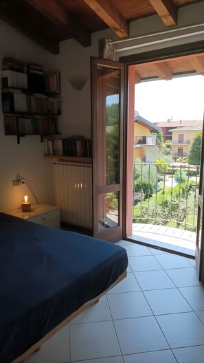 Une chambre à coucher comprenant un lit, une table de chevet, une lampe, une bibliothèque et une porte-fenêtre coulissante donnant sur un balcon avec vue sur le quartier.
