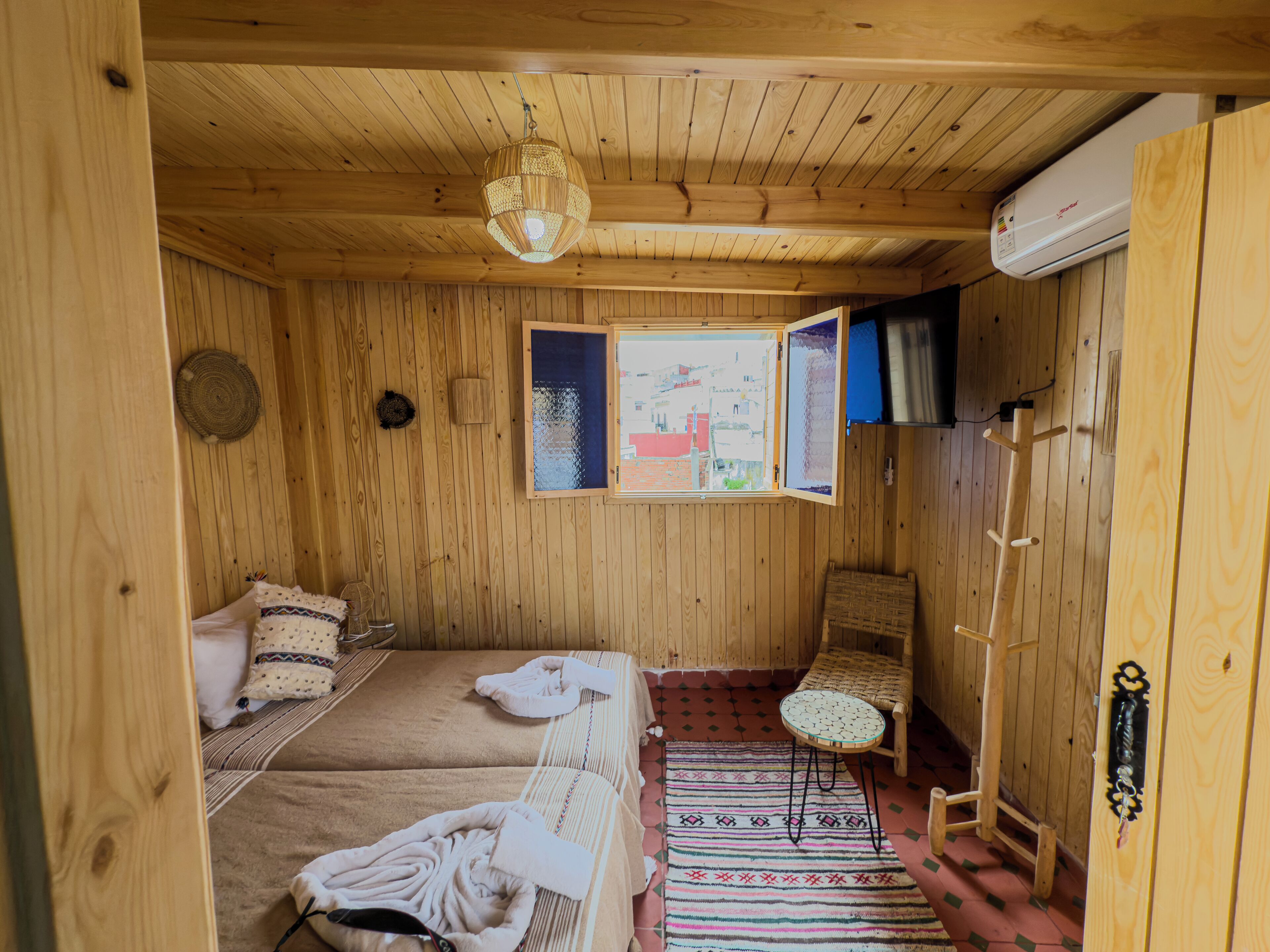 A wooden cabin room with two beds, a small table, a chair, a window with a view, and a hanging light fixture.