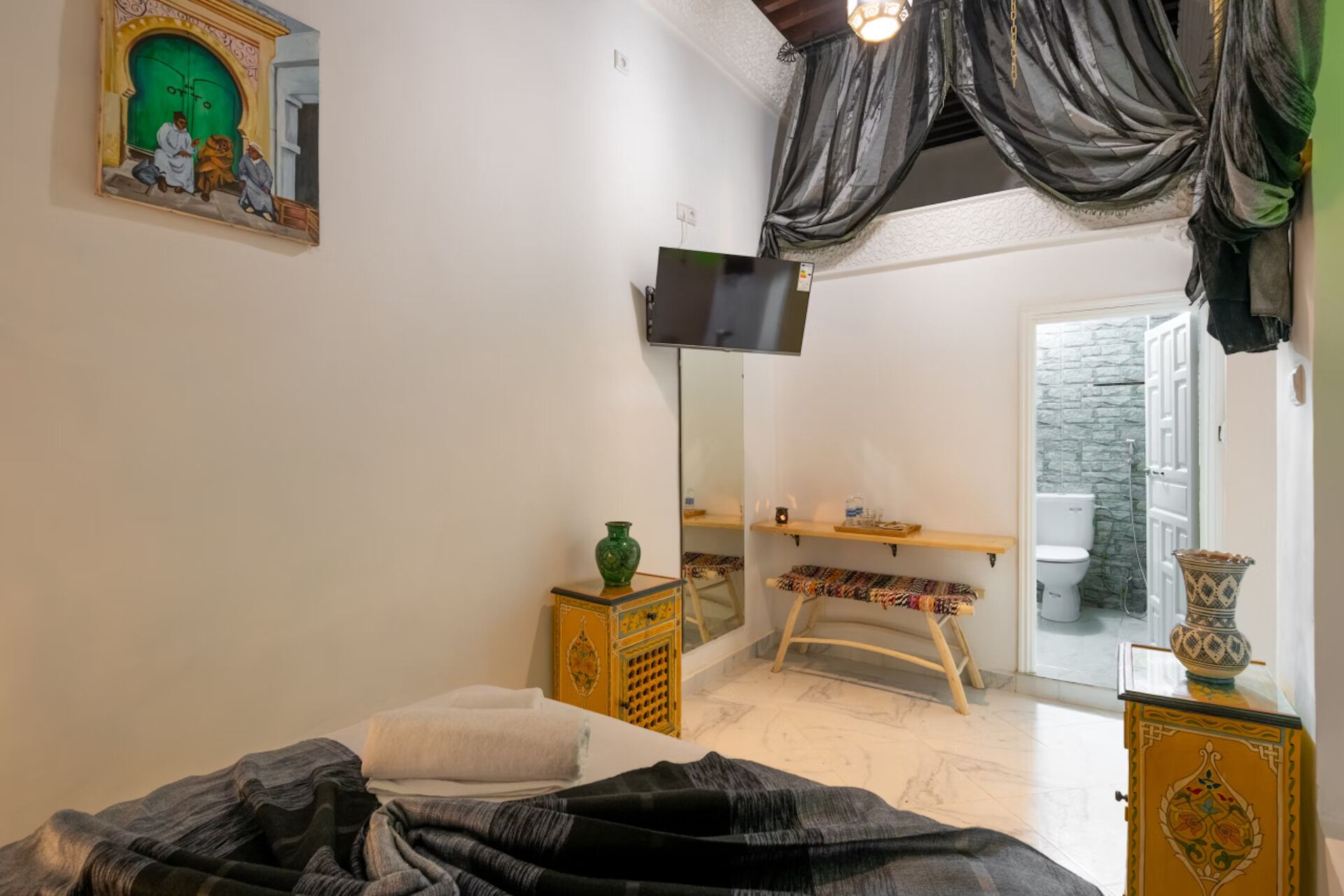 A bedroom with a bed, a mirror, a television, a small table with a vase, and a window with a view of a stone wall.