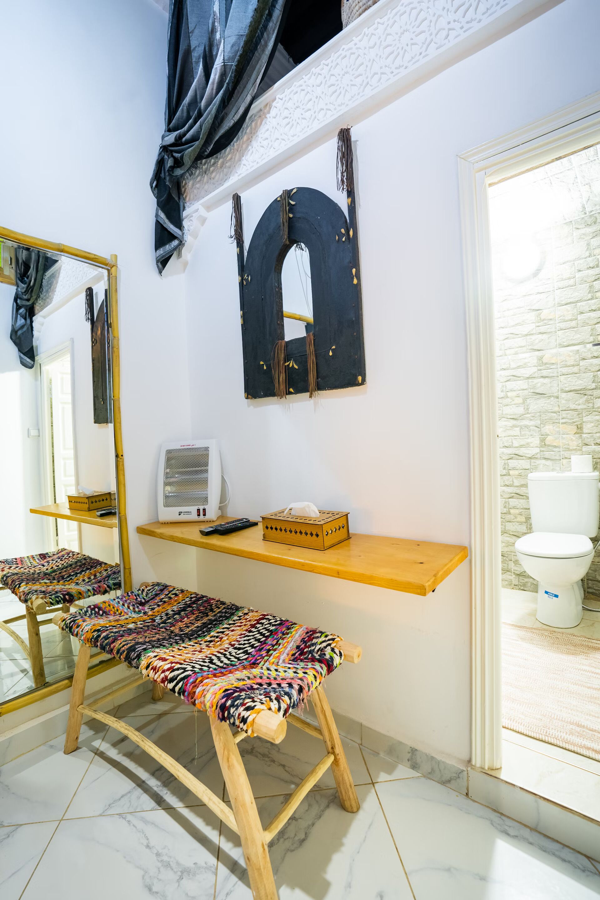 A bathroom with a white toilet, a wooden bench, a mirror, and a shelf with a speaker and tissues.