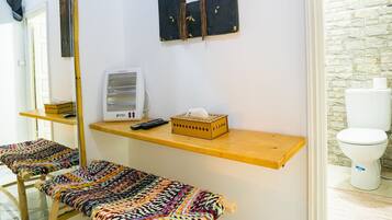 A bathroom with a white toilet, a wooden bench, a mirror, and a shelf with a speaker and tissues.