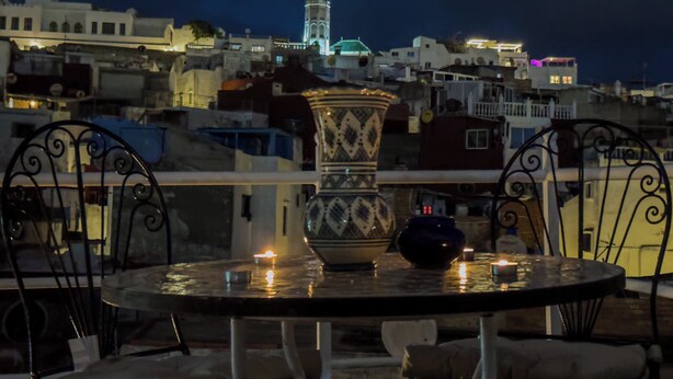 A rooftop terrace with a table and chairs, a decorative vase, and a view of a cityscape with illuminated buildings and a tower in the distance. - RIAD AMR (Tangier)