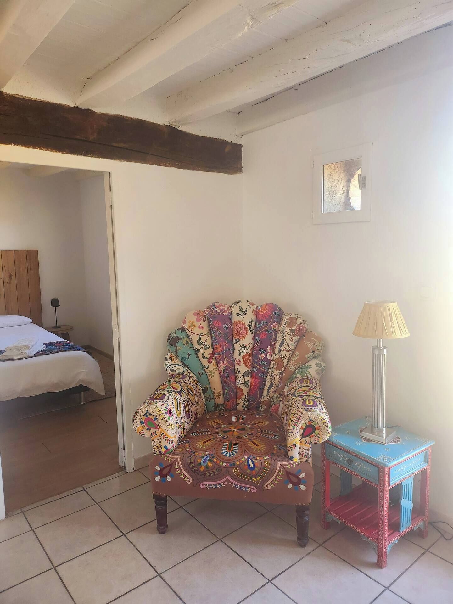 A room with a patterned armchair, a small table with a lamp, and a bed in the background.