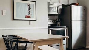 Una cocina moderna con armarios blancos, un refrigerador de acero inoxidable, un microondas y una estufa. Se observa un cuadro en la pared y una mesa de comedor de madera con sillas.
