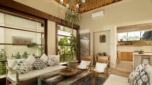 A cozy living room with a sofa, chairs, a coffee table, and a kitchen area visible through an open door. - Tranquil three bedrooms villa forest view in Jimbaran (Bali)
