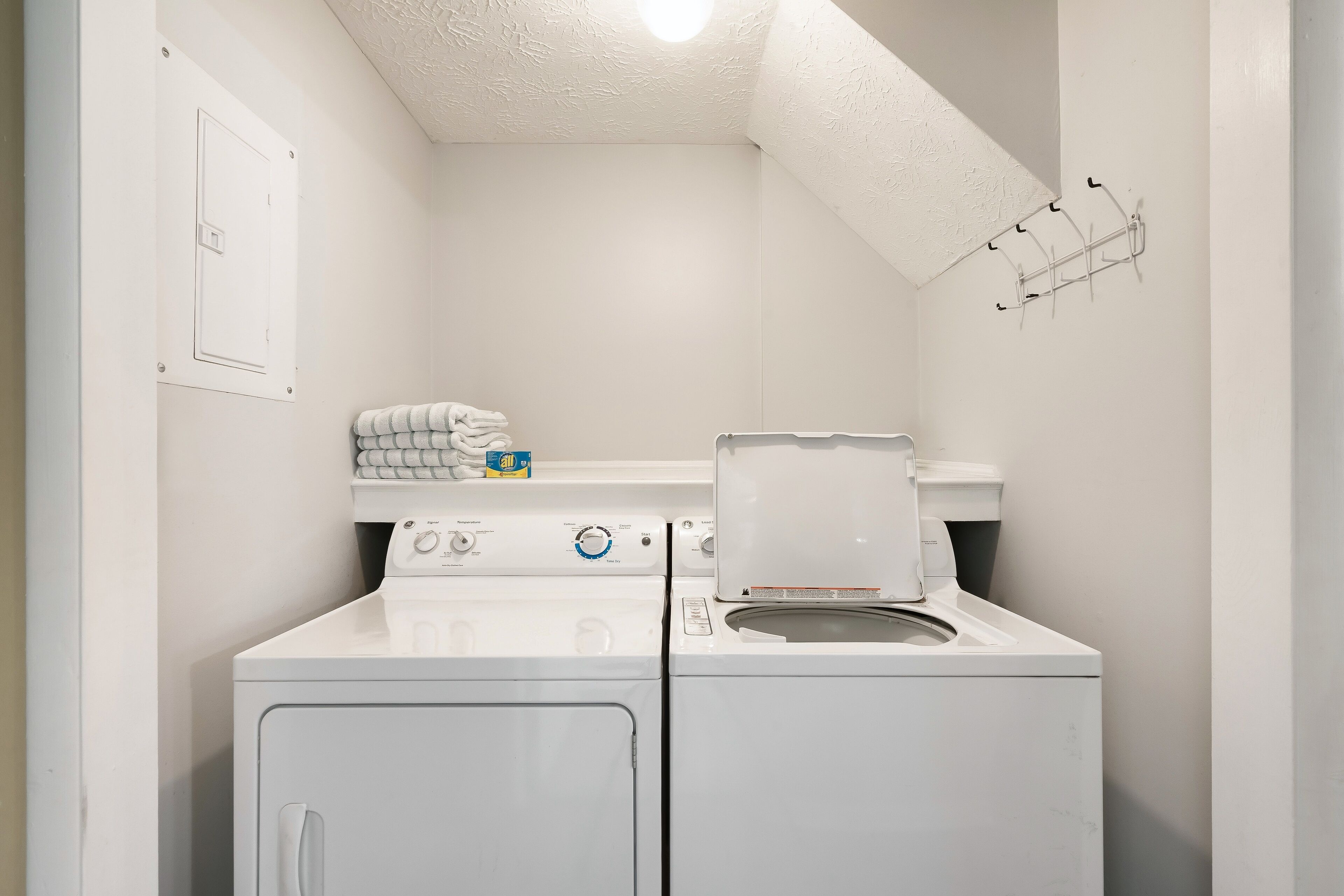 A compact laundry room with a front-loading washer and dryer, a towel rack, and a ceiling light fixture.