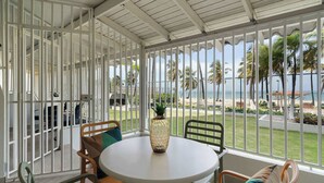 Een overdekt terras met een ronde tafel en stoelen, een decoratieve vaas, en uitzicht op het strand en palmbomen.