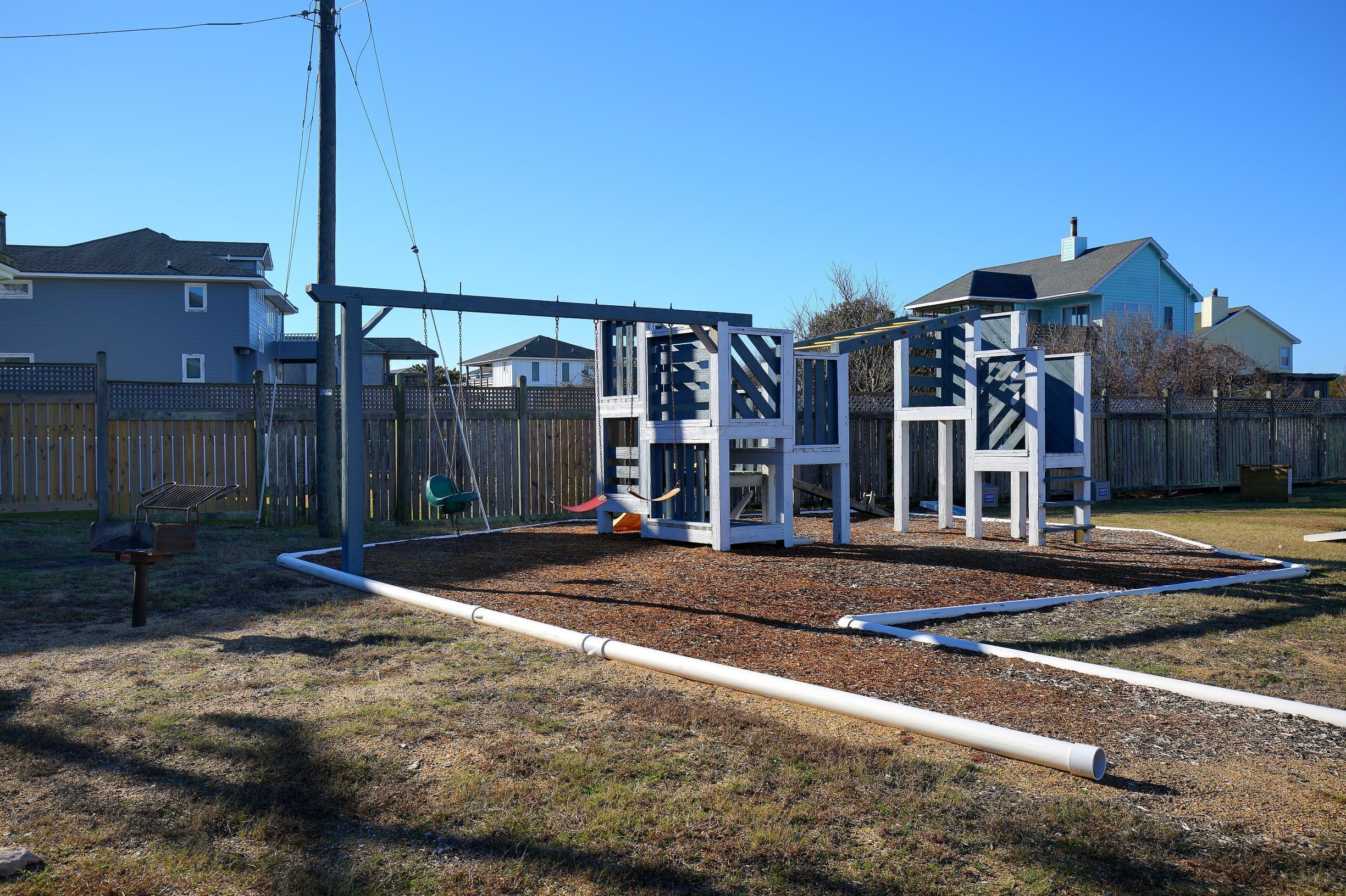 A playground with a swing set and slides, surrounded by a wooden fence and houses in the background.